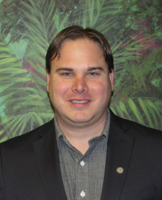 A man with short brown hair smiles, wearing a dark blazer over a collared shirt, against a vibrant green tropical background.